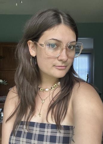 A young woman with long brown hair wears clear glasses, hoop earrings, layered necklaces, and a plaid strapless top, standing indoors in natural light.