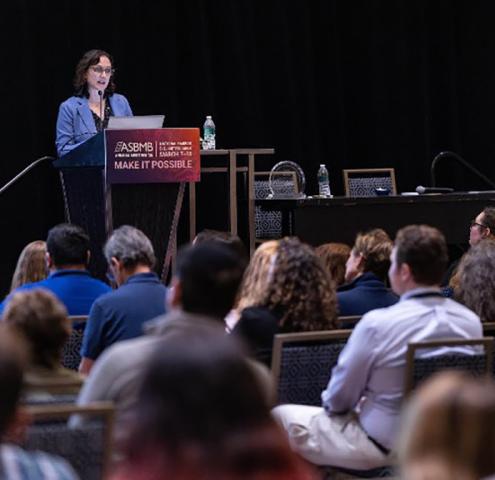 A woman speaks at a podium during a conference, addressing an audience seated in rows. A sign on the podium reads "ASBMB Make It Possible.