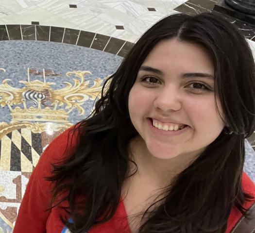A young woman with long dark hair smiles at the camera, standing indoors on a marble floor featuring a decorative crest design.