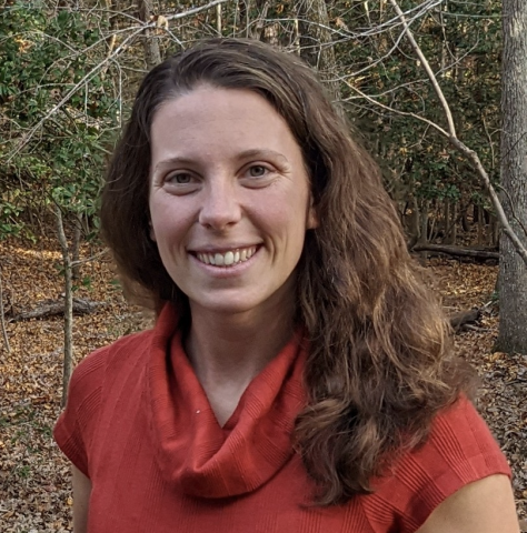 A woman with long brown hair, wearing a red cowl-neck top, stands outdoors in a wooded area with fallen leaves on the ground.
