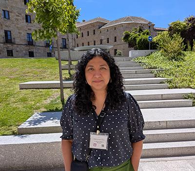 A woman with curly dark hair wearing a navy polka-dot blouse and name badge stands outdoors in front of stone steps and historic buildings on a sunny day.