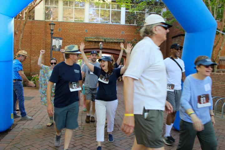 A group of people wearing numbered race bibs walk under a blue inflatable arch at an outdoor event; some are smiling and raising their arms.