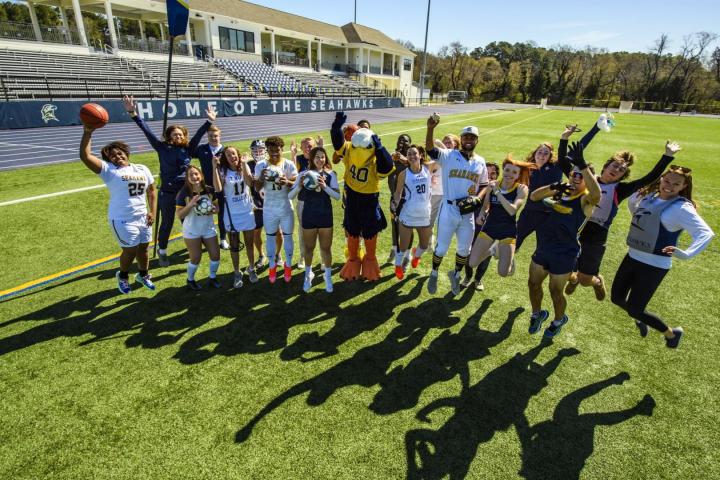 A group of athletes in sports uniforms and a mascot jump and pose for a group photo on a turf field in front of a stadium building.