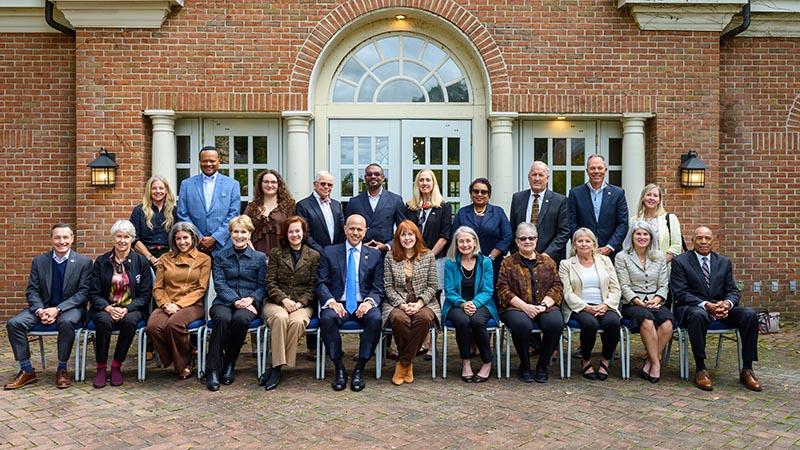 A group of 22 adults pose for a formal photo outside a brick building with arched windows, arranged in two rows—one seated and one standing.