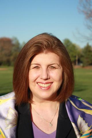 A woman with straight, shoulder-length brown hair smiles outdoors, wearing a black jacket and a colorful scarf, with trees and a blue sky in the background.