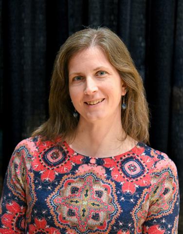A woman with long brown hair wearing a red and blue patterned top stands in front of a black curtain, smiling at the camera.