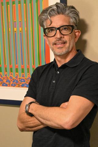 A man with gray hair, glasses, and a black polo shirt stands with arms crossed in front of a colorful abstract painting on a beige wall.