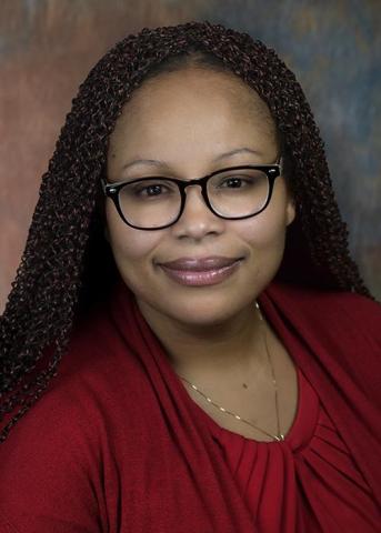 A woman with brown hair and glasses is wearing a red top, smiling at the camera against a neutral background.