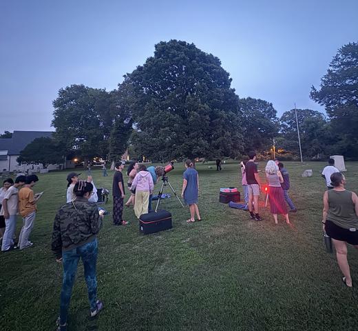A group of people gathers on a grassy field at dusk, some standing near telescopes, while others form smaller groups. Trees and buildings are in the background.