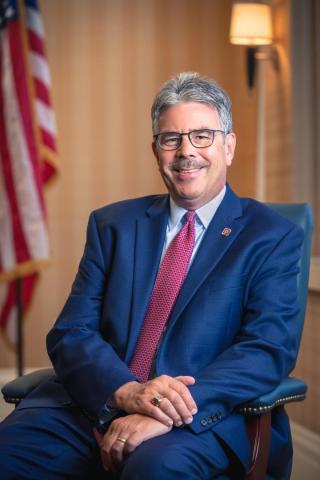 A man in a blue suit and red tie sits smiling in an office chair. An American flag and lamp are in the background.
