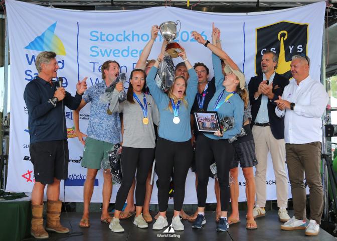 A group of people on stage celebrate with medals and a trophy at the World Sailing event, with banners and logos visible in the background. Photo by Malcom Hanes.