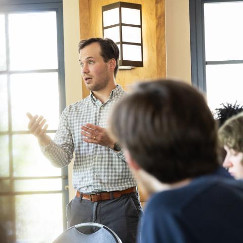 A man in a checkered shirt speaks while gesturing in a well-lit room, with two people seated in the foreground listening.