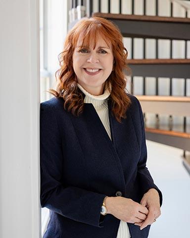 Woman with red hair wearing a navy blazer and white turtleneck stands smiling by a window, with shelves of binders in the background.
