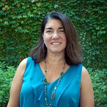 A woman with shoulder-length brown hair, wearing a teal sleeveless blouse and a long necklace, stands outside in front of green ivy and foliage.