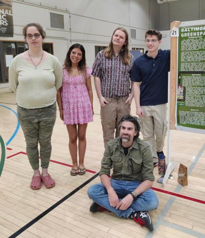 Five people pose by a science poster presentation in a gymnasium; four stand while one sits on the floor in front of the group.