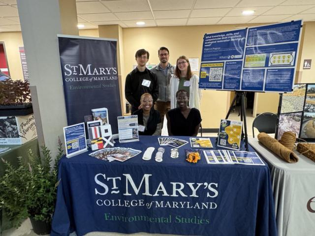 Five people pose at a St. Mary’s College of Maryland Environmental Studies booth featuring informational displays and brochures.