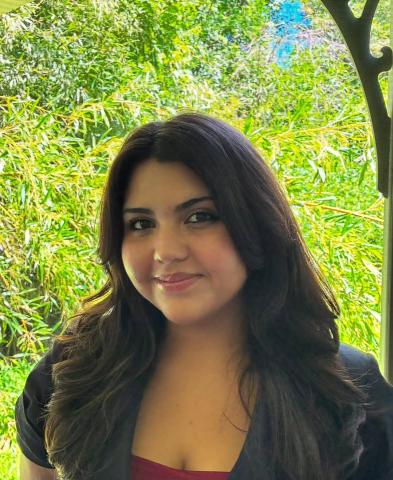 A woman with long dark hair and a black top stands in front of lush green foliage, smiling slightly at the camera.