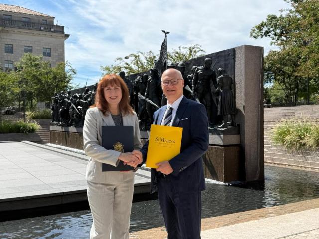 Two people in business attire shake hands outdoors in front of a large bronze monument, each holding official-looking folders.