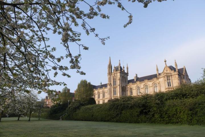 Large stone building with pointed towers sits behind a green lawn and blooming trees under a clear blue sky.