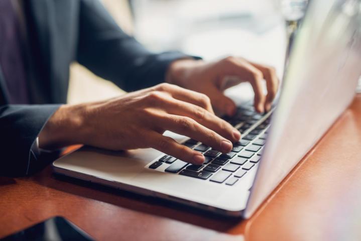 A person in business attire types on a laptop at a wooden desk, with part of a smartphone visible on the left side of the image.