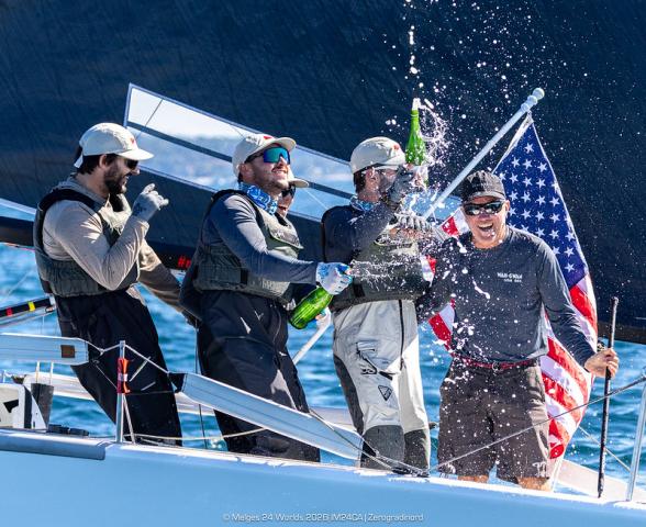 Four sailors celebrate on a sailboat, spraying champagne with an American flag in the background on a sunny day.