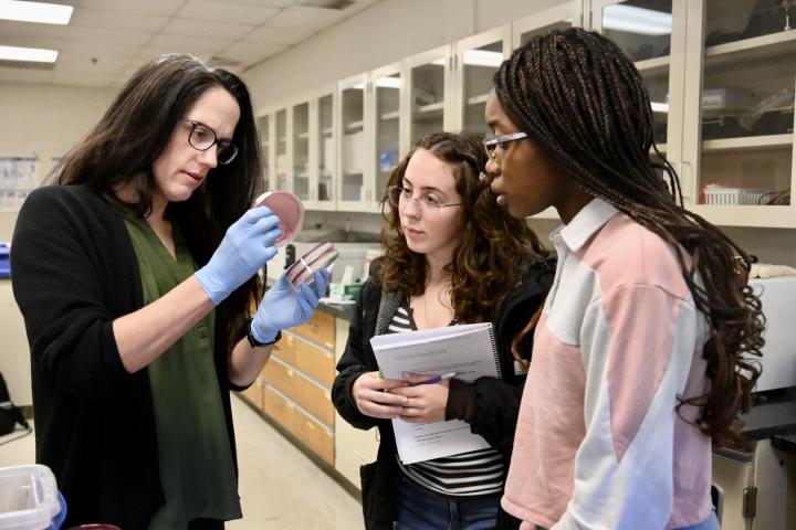 An instructor in a laboratory shows a petri dish to two students.