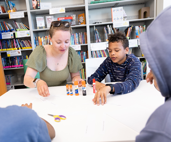 An adult and two children sit at a table working on a craft project with glue sticks and paper strips.