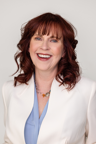 A smiling woman with shoulder-length reddish-brown hair wearing a light blue top and a white blazer.