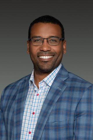 A smiling man wearing glasses and a plaid blazer over a light-colored button-up shirt.