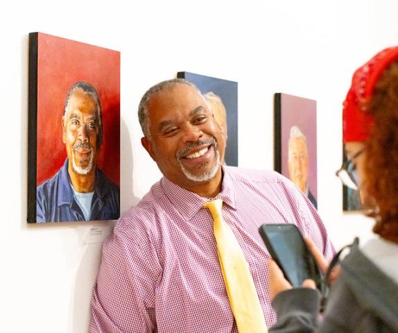 A smiling man in a pink button-down shirt and yellow tie stands in a gallery next to a portrait of a man, while a person in the foreground photographs him with a phone.
