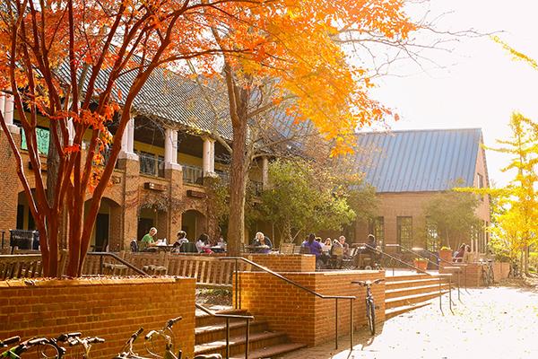 People sitting at tables on a brick patio in front of a brick building surrounded by autumn trees.