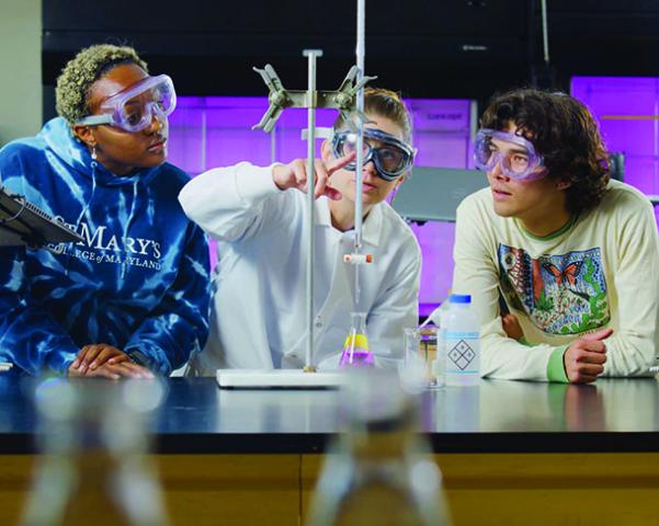 Three students wearing safety goggles observe a science experiment with beakers and lab equipment in a classroom setting.