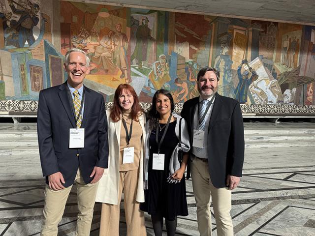 Four adults stand together outside in front of a mural wall, smiling at the camera.