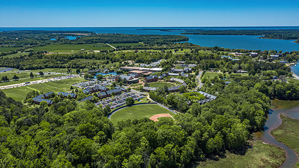 Aerial view of a campus with multiple buildings and a baseball field surrounded by trees, situated near a large body of water.