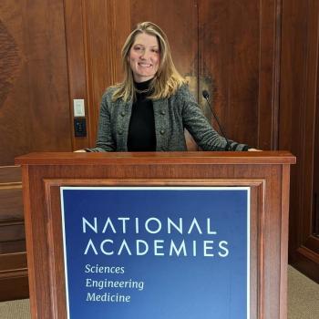 Neiles smiles while standing behind a podium labeled National Academies of Sciences Engineering and Medicine in a Grey blazer, black turtleneck, and gold necklace.