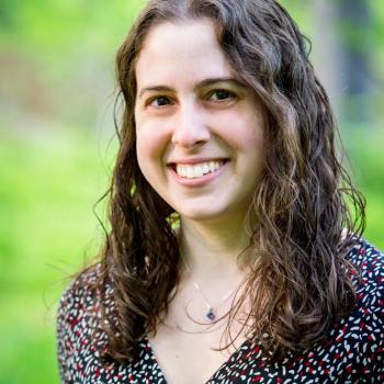 Dr. Gili Freedman, person with long brown curly harir smiling in front of a blurred image of greenery