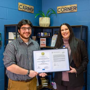 Two people are smiling and holding a framed certificate in a room with blue walls. Shelves with books and plants are in the background. Signs on the wall read "Caring Corner."