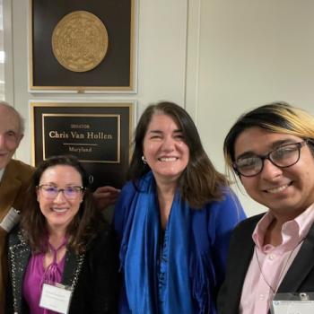 Four people are smiling and standing in front of a wall plaque for Senator Chris Van Hollen, Maryland. The group includes one person with glasses and another wearing a blue scarf. They appear to be in an office setting.