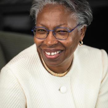 Smiling woman wearing glasses and a white textured blazer, sitting in front of a dark background. Her hair is short and gray, and she is accessorized with small gold hoop earrings.