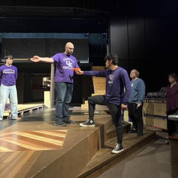 People on stage rehearsing. One person stands on a platform holding a script, while another person reaches out to them. Others watch nearby.
