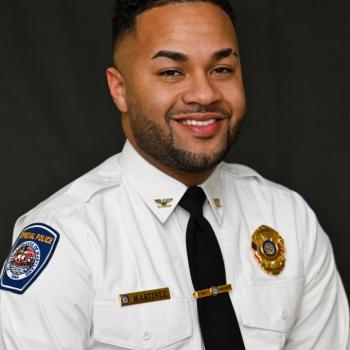 A man in a white police uniform with badges and a name tag that reads "Martinez" smiles against a dark backdrop.