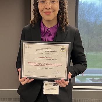 A person wearing glasses and a suit stands indoors holding a framed certificate and smiling at the camera.