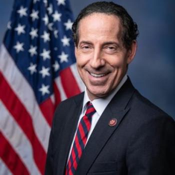 A man in a dark suit and striped tie stands in front of a U.S. flag, smiling at the camera against a blue background.