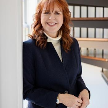 A woman with red hair, wearing a dark blazer and light turtleneck, stands indoors near a window, smiling with hands clasped. A spiral staircase is visible in the background.