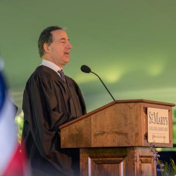 A man in academic robes speaks at a podium labeled "St. Mary’s College" during an outdoor event, with an American flag visible in the foreground.