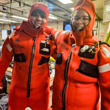 Two people wearing orange immersion survival suits smile inside a room filled with equipment and supplies.