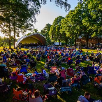 Large crowd sitting on lawn chairs and blankets at an outdoor concert in a park, with a stage set up under trees on a sunny day.
