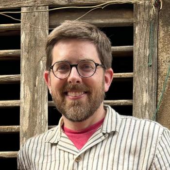 A bearded man with glasses and short hair, wearing a striped shirt and red t-shirt, stands in front of a weathered wooden window.
