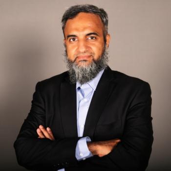 Man with a gray beard wearing a black suit jacket and light blue shirt, standing with arms crossed against a plain gray background.
