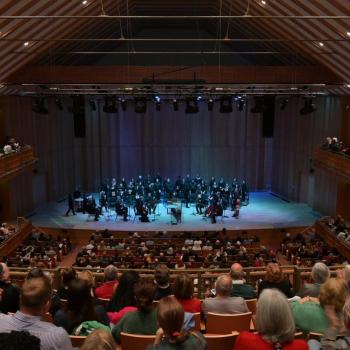 A symphony orchestra performs on stage in a wooden concert hall, with audience members seated on the main floor and balconies.